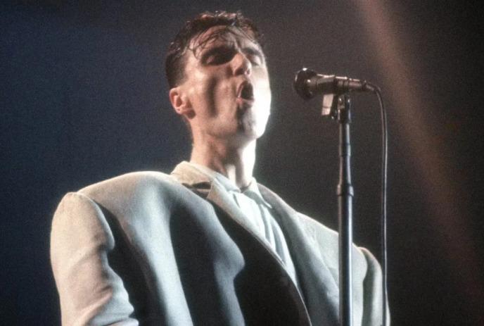 Performer in oversized light suit stands at microphone under spotlight against dark stage background.