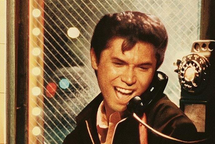 Person holding black rotary phone near mouth with short hair jacket and colorful lights behind.