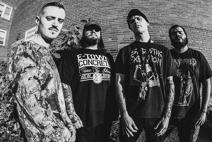 Four men of the band The Last Ten Seconds Of Life stand in front of a brick wall, wearing dark graphic tees.