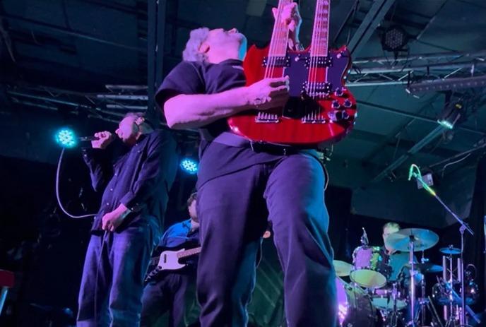 Musician plays red double-neck guitar on stage as singer performs nearby, lit by dramatic blue and purple concert lights.
