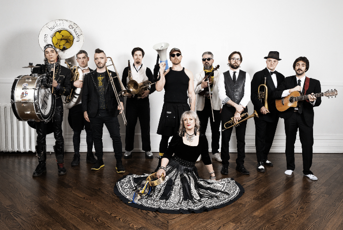 Ten musicians pose indoors with varied brass and string instruments against plain white wall backdrop setting around.
