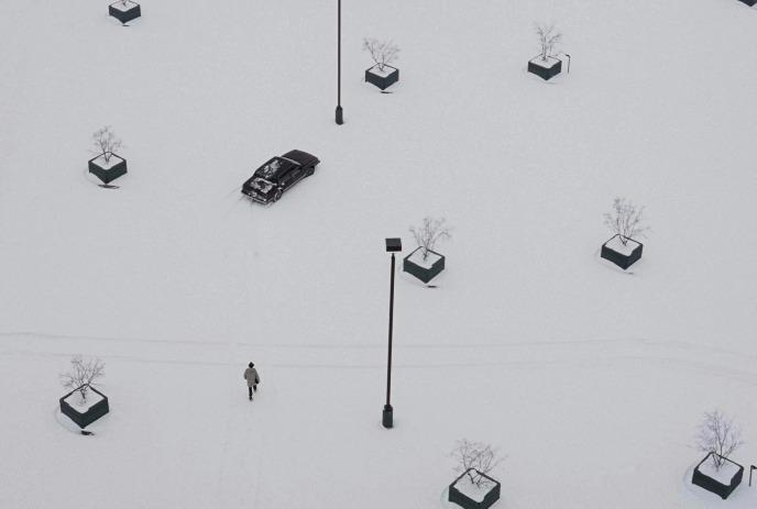 Aerial view of a snowy plaza with scattered bare trees in planters, one pedestrian walking and a black car parked in the snow.