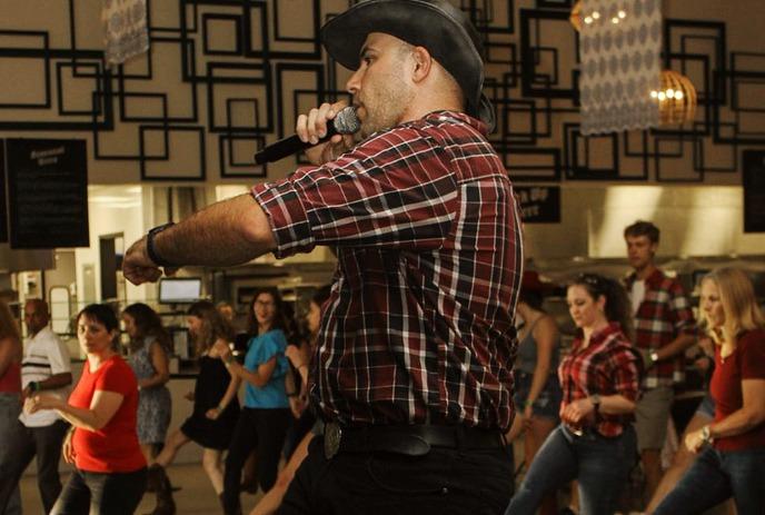 Man in cowboy hat and plaid shirt holds mic mid-dance, warm indoor crowd watches under hanging lights.