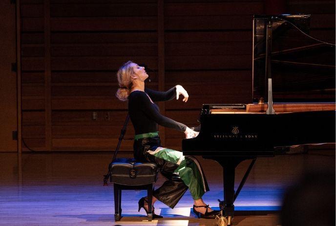 Pianist performing on grand piano in wood stage hall wearing black top and green pants focused on playing.