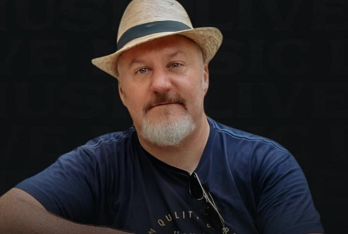 Artist in straw hat and dark shirt resting arm on guitar before dim backdrop with subtle live music theme.