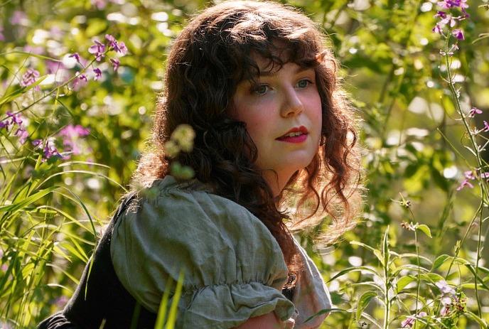 Woman with curly hair sits in a lush green field surrounded by wildflowers.