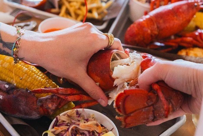 Two hands pull apart cooked lobster, revealing white meat; corn, coleslaw, and fries surround seafood feast.