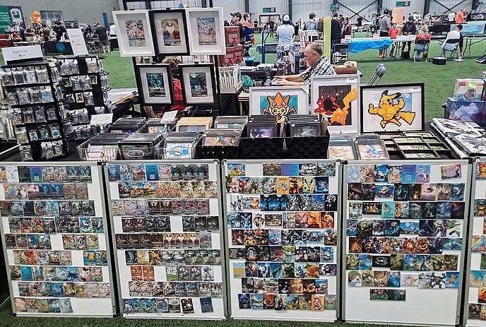 Vendor booth with colorful trading cards, framed art, and people browsing at indoor convention.