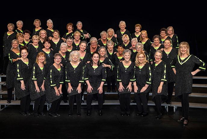 A group photo of the London Chorus, featuring women in sparkling black outfits with neon green accents, posed in rows on a stage.