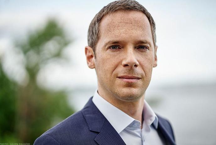 Man in blue suit by water, trees and sky behind.