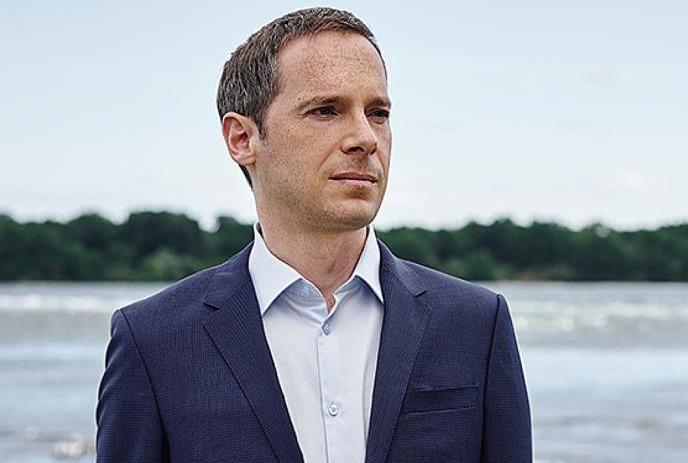 Artist in blue suit stands by water with trees in background with clear sky.