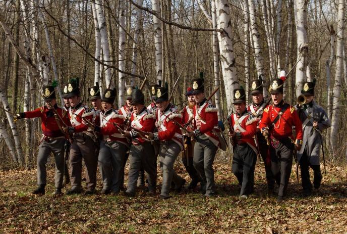 A group of people dressed in historical red military uniforms with black shako hats march through a forest.