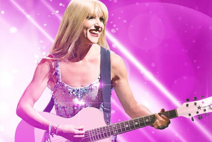 A smiling Taylor Swift impersonator with long blonde hair plays a pink guitar on stage under colorful blue and pink lights.
