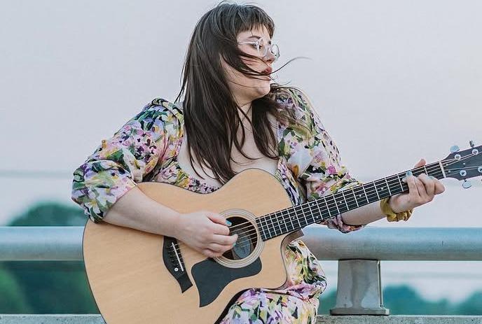 Artist sits on concrete ledge playing guitar as wind lifts hair against soft outdoor sky backdrop.