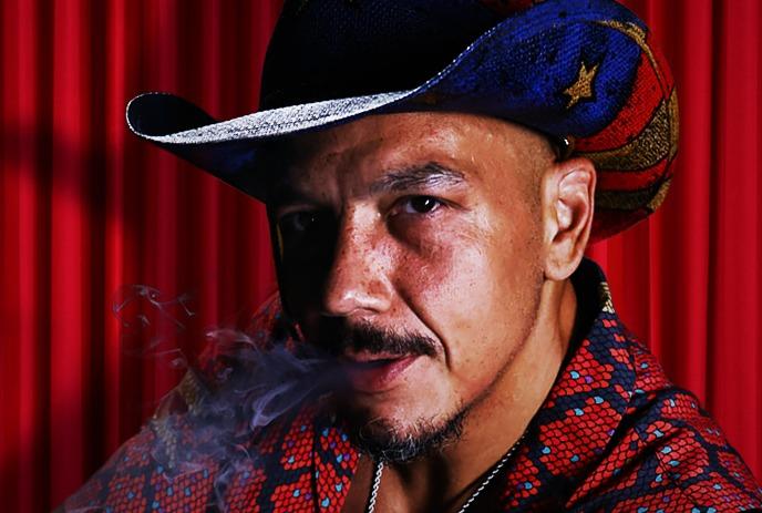 Luis J Gomez with a mustache in a red patterned shirt and a star-spangled cowboy hat smokes against a vibrant red curtain.