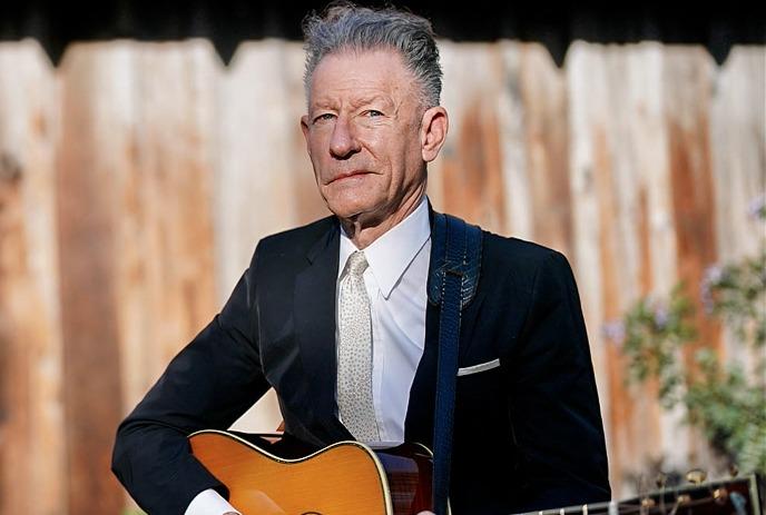Lyle Lovett standing outside in front of a wooden fence, wearing a suit and holding a guitar.