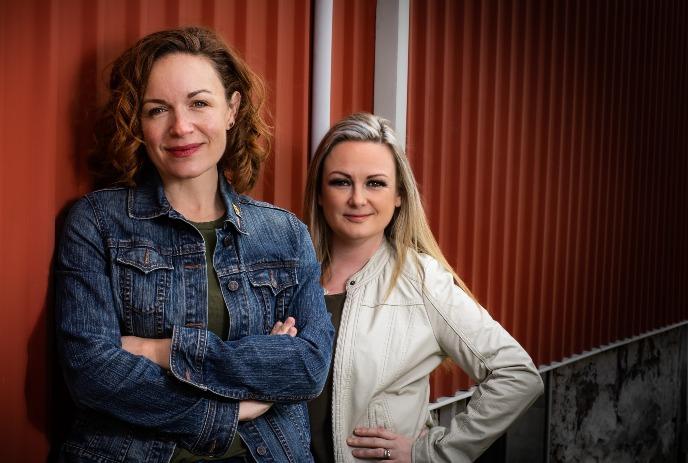 Two artists posing confidently against corrugated red metal wall wearing casual jackets outdoors together.