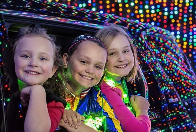 Two children in colorful winter hats pose near a glowing Magic Lights display at night.