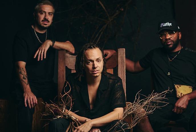 Three musicians pose with natural branches against a colorful teal and purple graphic backdrop.