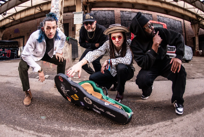 Four musicians pose around an open guitar case beneath an overpass.