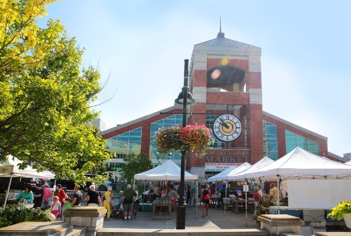 Sunny outdoor farmers' market at Covent Garden with white tents, shoppers, flowers, and a historic brick building.