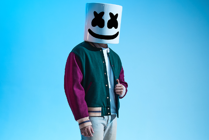 Musician Marshmello poses against a blue background, wearing his signature white helmet and a varsity jacket.
