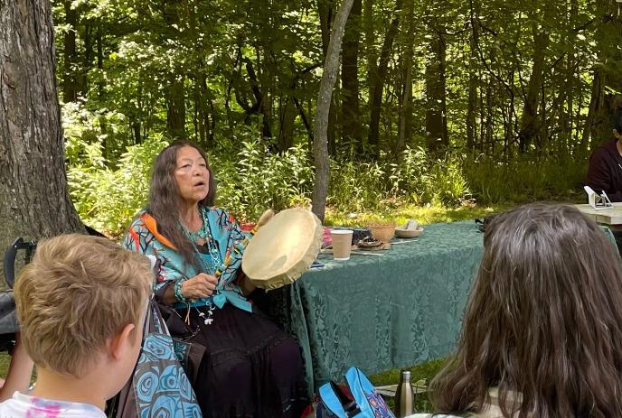 Person in colorful attire plays hand drum outdoors while small seated group listens amid trees and natural setting.