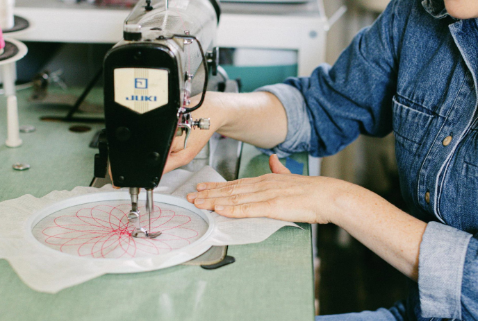 Person uses sewing machine to stitch red circular pattern onto white fabric held in hoop with careful guidance.