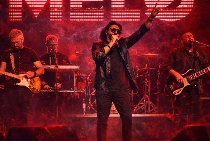 Band performs on red lit stage as singer raises arm with guitarists flanking and drums behind.