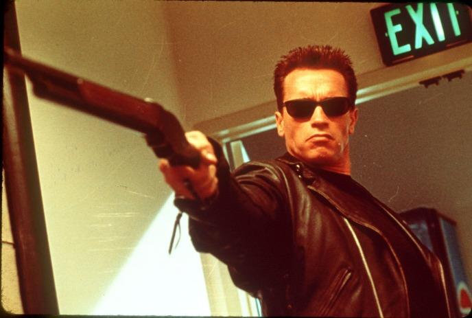 Person wearing all black and black sunglasses, pointing a gun at something out of sight.