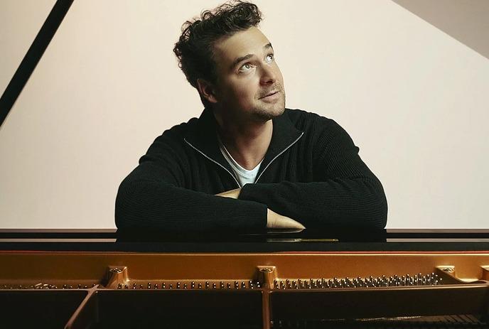 Person seated at grand piano rests arms on edge while gazing upward against softly lit calm background setting.