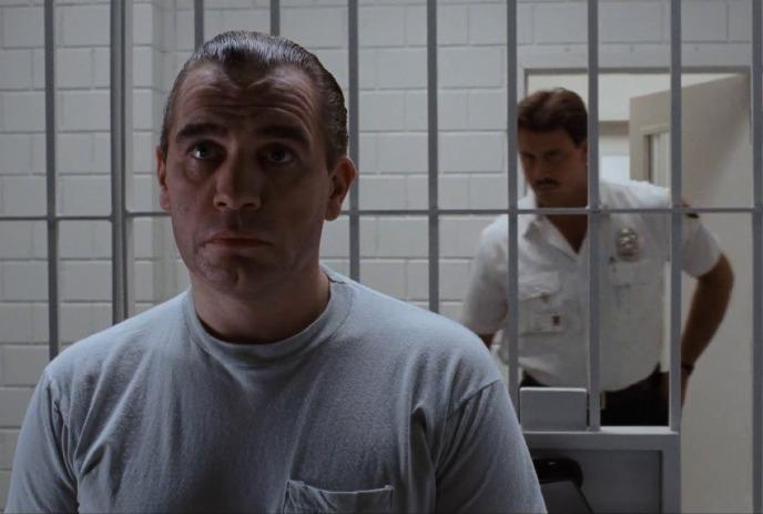 Person standing before metal jail bars with guard visible behind in tiled room creating tense confined scene.