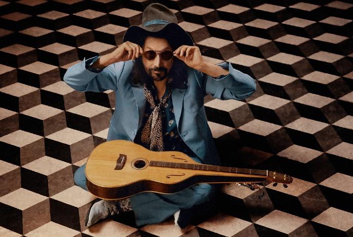 Musician Mike Campbell sitting on a geometric patterned floor wearing a blue suit.