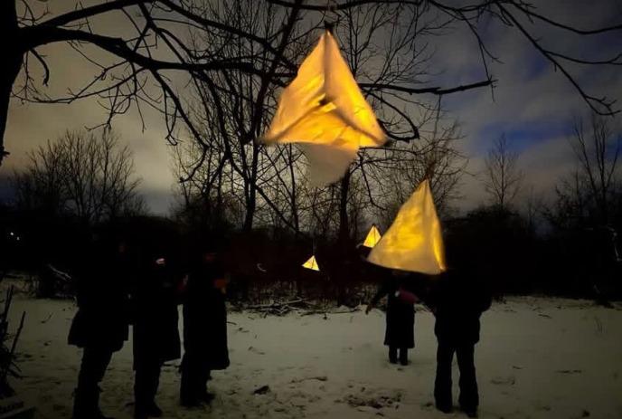 A snowy outdoor scene with people standing under glowing yellow triangular lanterns hanging from bare trees.