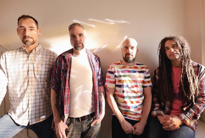 Four artists dressed stand side by side, each in distinct shirts: plaid, checkered, striped, posing for a picture.