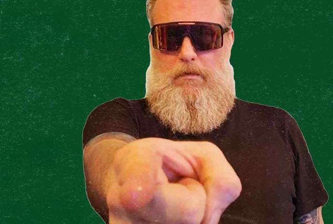 Man with white beard and dark sunglasses points at camera against solid green background; assertive, bold gesture.