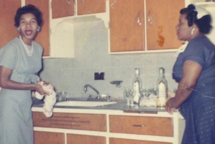 Two women in vintage-style blue dresses stand in retro kitchen with wooden cabinets.