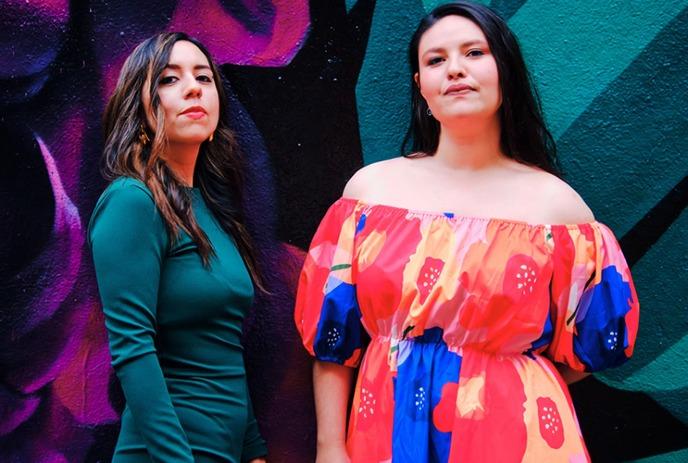 Two women pose in front of a vivid mural; one wears green, the other a floral dress with bold colors.