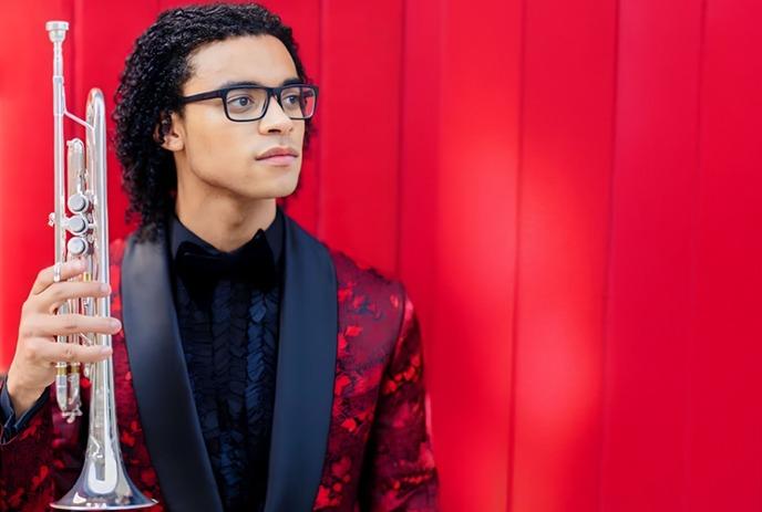 Will Leathers holding a trumpet, dressed up wearing glasses, in front of a red background.