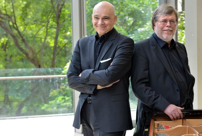 Leslie Kinton and James Anagnoson wearing black suites, posing for the camera in front of trees on a balcony.
