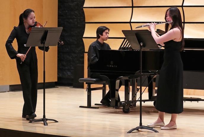 Three musicians perform on stage: a violinist, flutist, and pianist, each reading sheet music in a warm wooden concert hall.