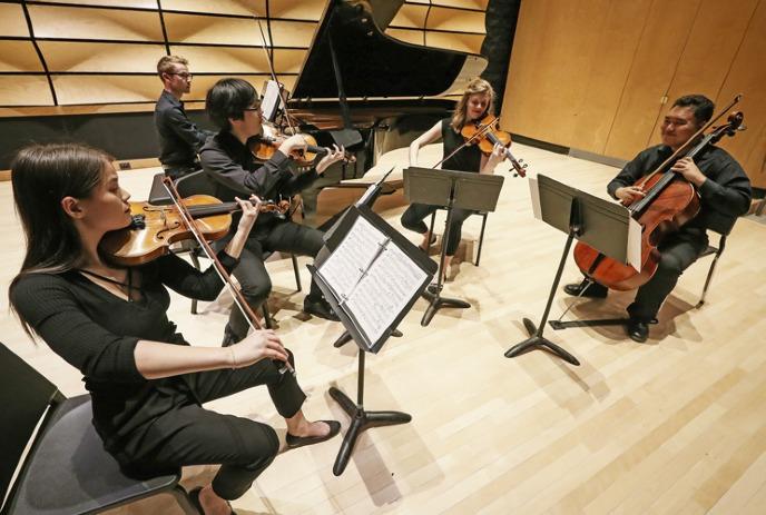 Musicians in black play violins and cello on stage with sheet music and a piano.