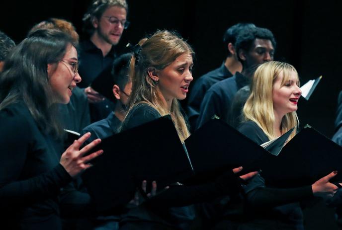 A diverse choir of young adults singing passionately with open music folders.