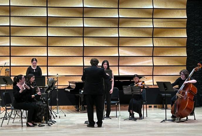 Musicians in black outfits performing on stage with conductor, wave-pattern wood wall behind.
