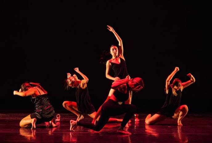 Five dancers in black perform on stage under dramatic red lighting.