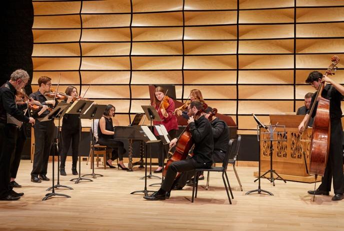 A chamber orchestra performs on a wooden stage with a wave-patterned backdrop.