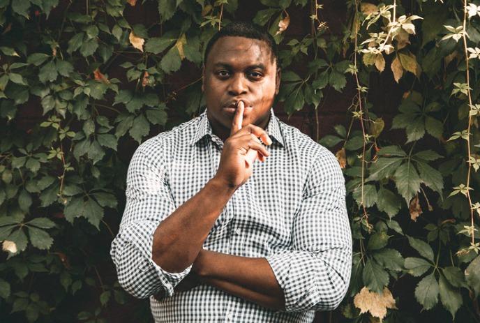 A man in a checkered shirt stands in front of lush green ivy, holding a finger to his lips in a 