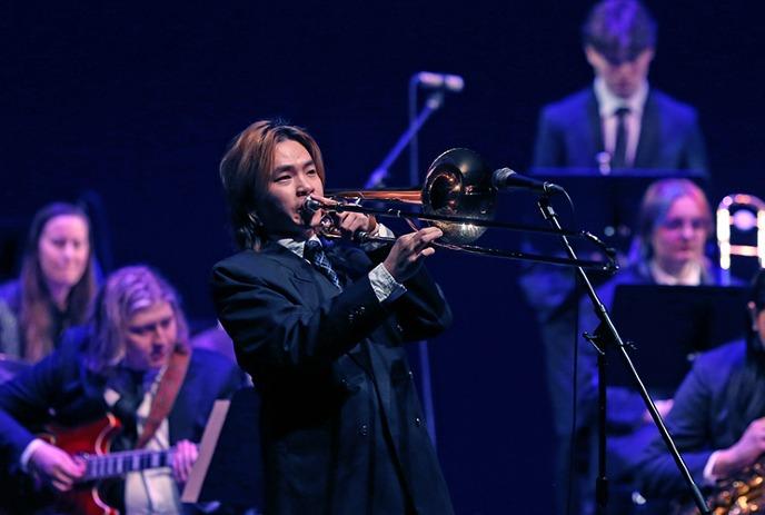Jazz ensemble performs on stage: trombonist in foreground with guitarist and saxophonist in suits under blue lights.