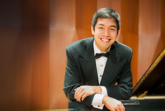 Dan Luong, a pianist in a tuxedo, smiling and leaning on a grand piano.