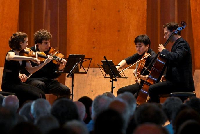 String quartet performs on stage; focused musicians play to attentive audience in warm, wood-paneled hall.